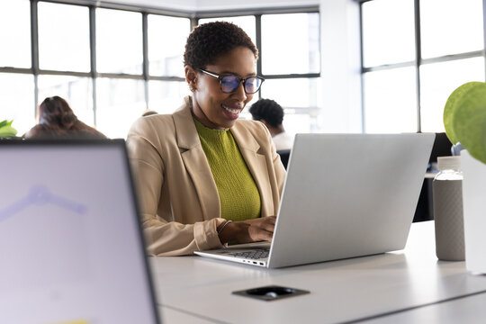 African American adult woman smiling, typing on silver laptop in office in beige blazer and glasses