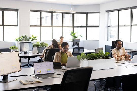 Diverse coworkers working in open office in business-casual, viewing charts on laptops and monitors