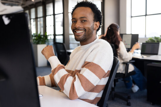 African American man wearing striped sweater turning to camera smiling at office desk with monitor