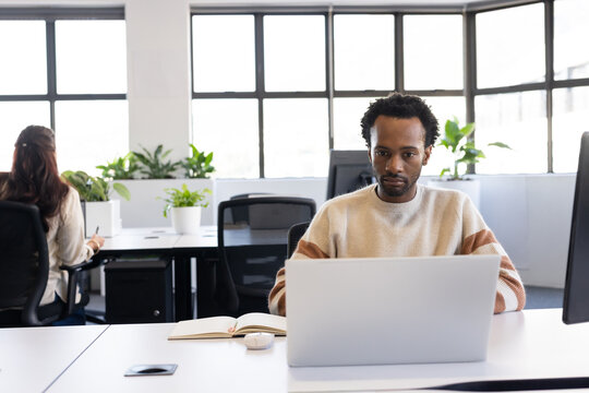 Diverse coworkers with African American man working on silver laptop in open-plan office, plants
