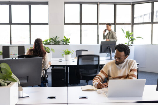 Diverse coworkers working in bright office, desk man writing in notebook near laptop, copy space