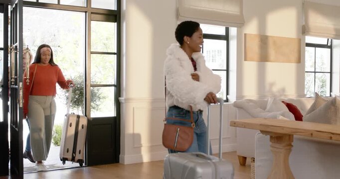 Diverse friends entering glass door led by woman in white jacket pulling luggage for settling in