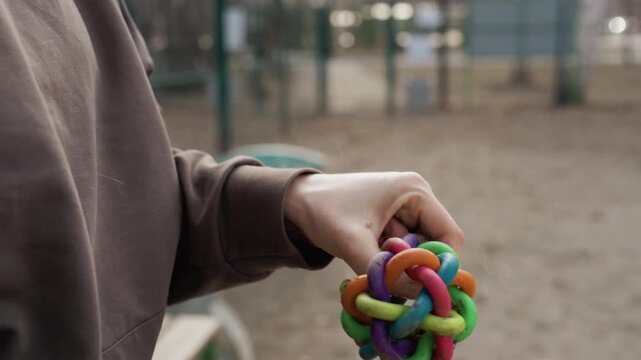 Person manipulates colorful interwoven plastic toy in outdoor setting. person hands and vibrant object are shown interacting against blurred background of playground equipment and open space