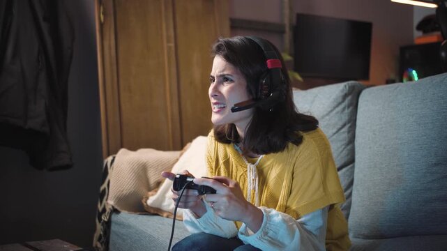 Young Latina woman sitting on her sofa at night, playing console video games and talking with friends through her gaming headset, representing online gaming and social connection.
