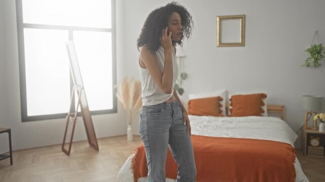 Woman talking on smartphone and gesturing by bed in a sunlit bedroom with orange bedding and mirror; deciding outfit thoughtful.