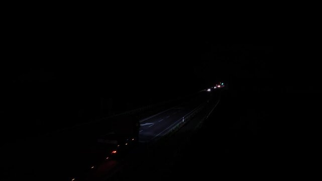 Truck with red taillights driving on dark highway at night, oncoming cars with bright headlights, white lane markings in USA, glowing vehicle lights in deep darkness. Aerial drone view.