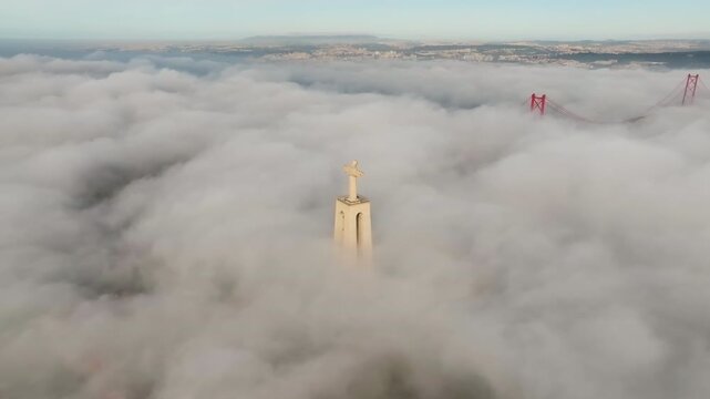 Slow drone pan of Cristo Rei at sunrise, Lisbon. Dense fog covers water. Christ the King statue and upper 25 de Abril Bridge emerge. Portugal aerial footage