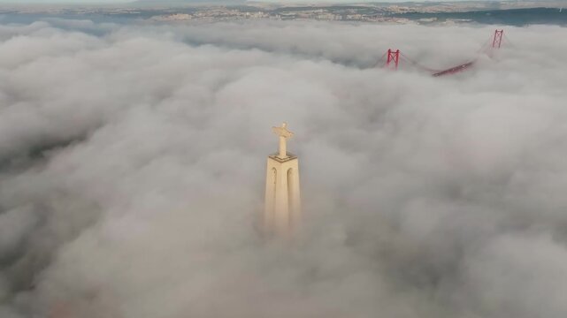 Slow panning drone shot of Cristo Rei, Lisbon. Dense fog blankets water at sunrise. Christ the King and 25 de Abril Bridge top visible. Portugal footage