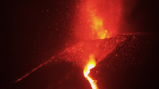 Molten rock cascades down a dark cinder slope while incandescent debris ejects into the night sky.