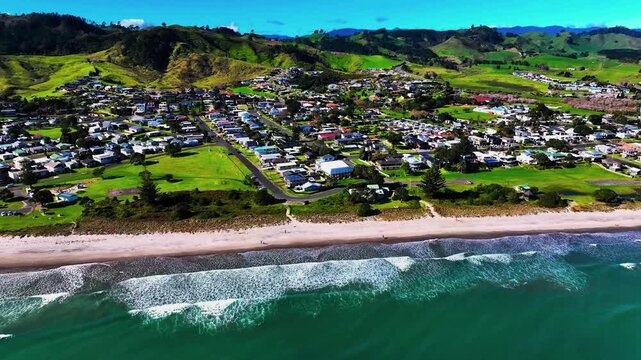 Waves break along a long golden shoreline beside green hillside settlements and rolling pastures.