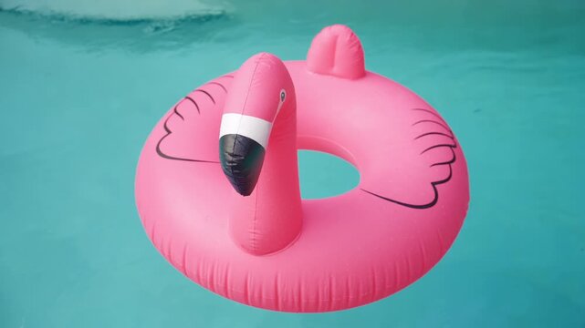 A pink inflatable flamingo ring floats on turquoise swimming pool water. Water drops on the vinyl, black beak and wing drawings. Summer vacation fun, relaxing holiday mood, gentle ripples.