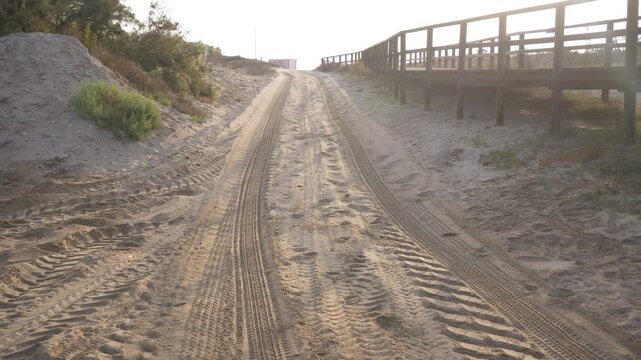 Tire tracks from a tractor and 4x4 on a sandy path heading toward coastal dunes, with a wooden boardwalk running parallel on the right. Natural outdoor scene, sunny day, beach environment.