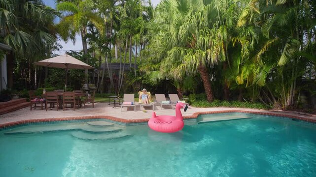Beautiful girl relaxing by the pool on a sun lounger in a hat. Pink flamingo float on turquoise water. Tropical backyard with palm trees, patio umbrella and dining set. Sunny summer vacation.