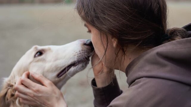 Woman wearing brown hoodie interacts affectionately with light colored dog gently petting and nuzzling it in outdoor setting. scene focuses on close up details of their interaction