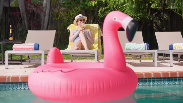 Beautiful girl relaxes by the pool on a sun lounger with a yellow lemon towel, smiling and raising a cocktail glass. Pink flamingo float in blue water. Sunny summer vacation among tropical palms.