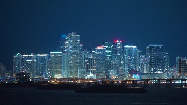 Red aviation lights crown glass towers of downtown Night Miami. Illuminated skyline over dark Biscayne Bay. Causeway bridge with car lights and a small island in front. Deep blue night sky.
