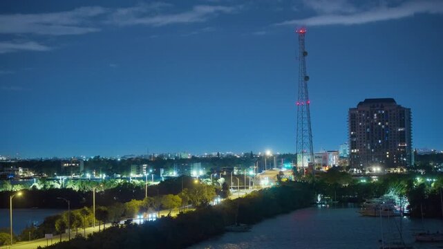 A tall lattice cell tower rises over a lit causeway road with car headlights in night Miami. Marina boats, palm trees, a residential high-rise and city lights glow under a deep blue evening sky.