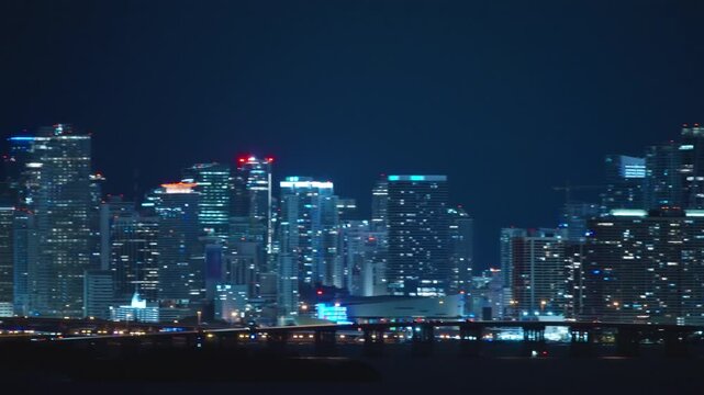 Night Miami skyline of illuminated glass skyscrapers seen across dark Biscayne Bay. Causeway bridge with car lights, red aviation beacons, construction crane. Downtown and Brickell towers.