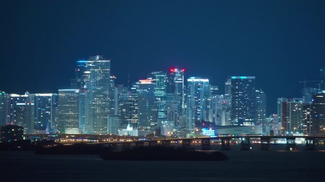 Night Miami skyline panorama from Brickell to downtown with a lit causeway bridge on dark piers. Glass skyscrapers with glowing windows, a construction crane and palm trees on the calm bay shore.