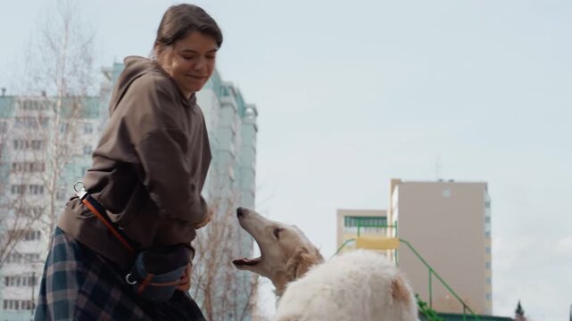 Woman interacts with dog in outdoor urban setting holding and throwing ball while white coated animal eagerly follows. scene features tall apartment buildings bare trees soft daylight