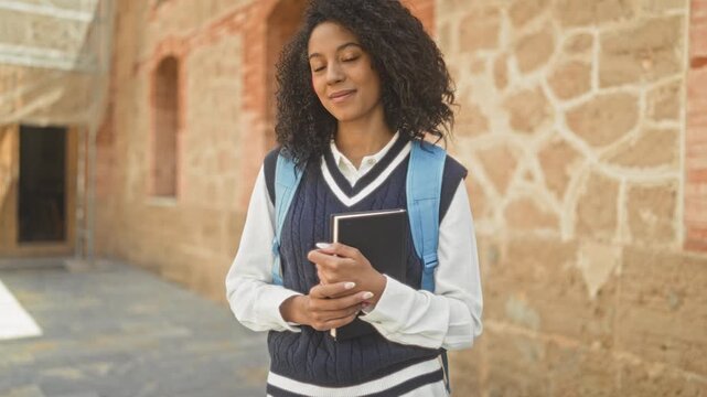 Young woman student holding notebook and blue backpack, smiling with eyes closed and hands clasped in front of stone building; optimism study focus.