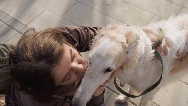 Woman interacts affectionately with white dog on outdoor paved area. Woman pets and kisses calm relaxed animal while wearing casual clothing and carrying backpack. scene features soft