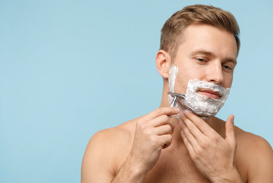 Shirtless man applying shaving cream and using a double edge razor on his cheek, morning grooming routine and personal hygiene isolated on light blue