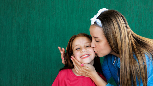 Happy latin mother having tender moment kissing daughter on green background - Family love and motherhood concept - Focus on child girl face