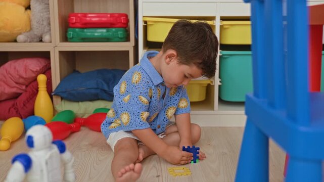 Boy sitting crosslegged on floor assembling colorful interlocking toy pieces beside storage shelves inside a building; concentration curiosity.