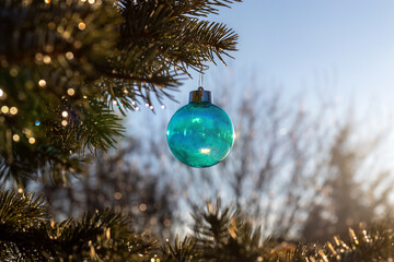 Obraz premium Turquoise Christmas bauble hanging from spruce branch outdoors. Christmas holiday decoration with golden reflections. Seasonal celebration and winter greeting.