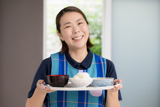 A smiling care staff member serving a meal; a home health aide providing meals as part of in-home care or housekeeping services, looking directly at the camera