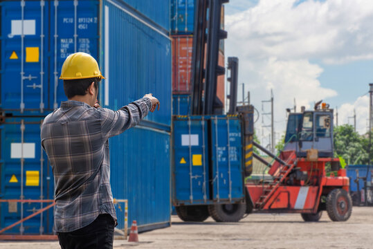 Logistics manager wearing safety helmet directing operations at cargo terminal with shipping containers and reach stacker. Global supply chain concept. Human-created, not AI.