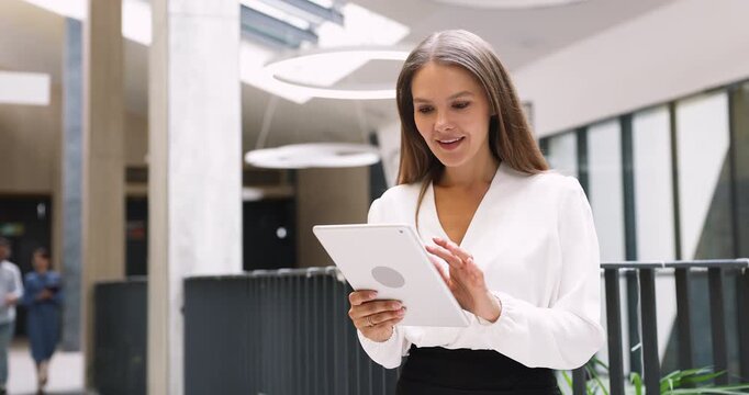Female professional checking corporate app updates on digital tablet in atrium office walkway reviewing completed tasks enjoying positive work results receiving good news staying connected with smile