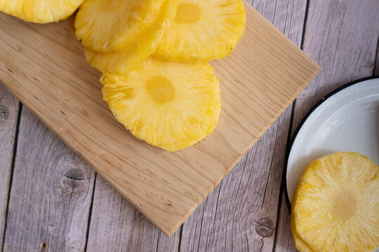 Fresh pineapple slices rest on wooden cutting board. Slices display vibrant yellow flesh and juicy texture. Plate holds extra slice nearby