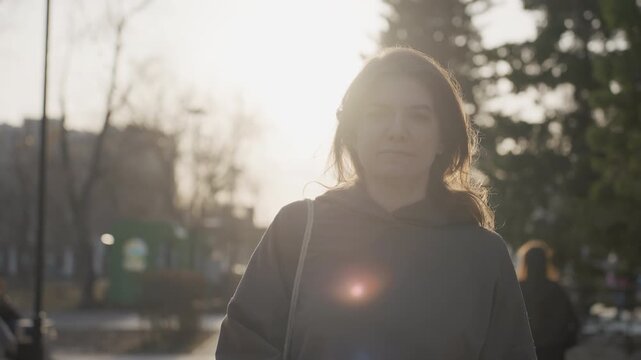 Woman stands outdoors in park during sunset wearing dark hoodie with sunlight backlighting her hair and shoulders. background shows blurred trees distant buildings other people walking