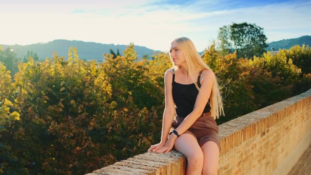 A blonde woman in a black tank top and brown skirt sits on a stone parapet wall, gazing into the distance at a scenic overlook with lush hills and greenery.