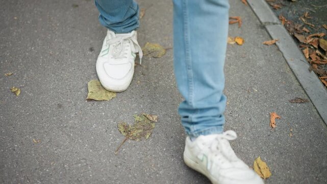 Close up of person feet in white sneakers and blue jeans walking on pavement with scattered autumn leaves. focus remains steady capturing movement from various angles as they walk forward
