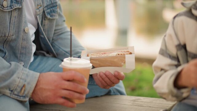 Close up of two people sitting at wooden table outdoors one holding coffee and sandwich while other holds food. scene features casual interaction with visible denim jackets and striped
