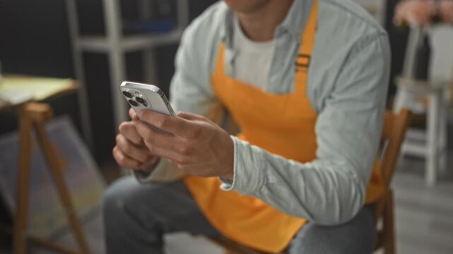 Young painter man in orange apron seated holds smartphone with both hands, scrolling and looking down at the screen in an art studio with easels and shelves; concentration.