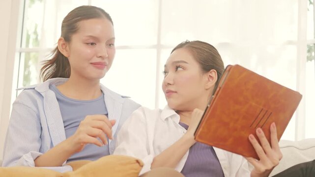Happy young lesbian couple relaxing on a couch, browsing the internet on a tablet, and talking happily.	