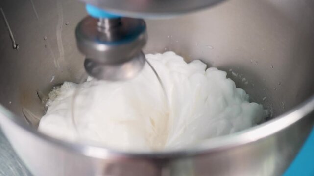 Close-up of electric mixer whisk beating thick white cream in stainless steel bowl. Smooth texture, professional pastry kitchen, baking preparation. Slow motion Full HD video