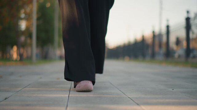 Close up of person feet in pink shoes walking steadily on outdoor paved path with soft lighting. subject wears black pants and background shows blurred trees and distant structures