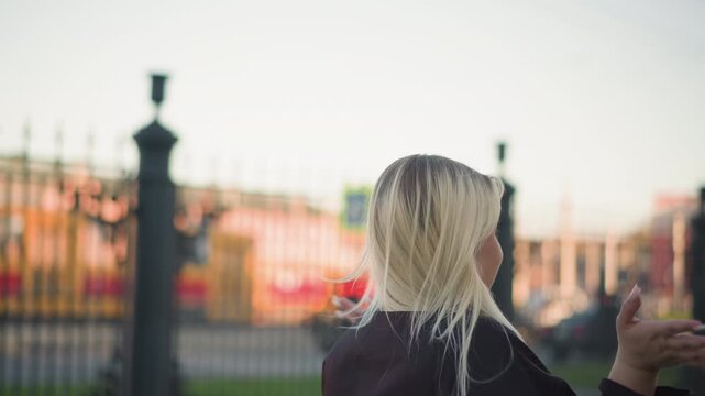 Close up of woman with long blonde hair wearing black standing outdoors facing away from camera. Her posture remains mostly still with subtle head and arm movements visible in later frames