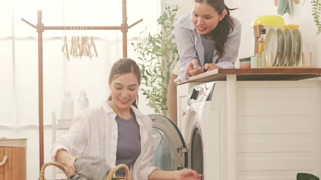 Cheerful Asian lesbian couple doing laundry together at home, loading the washing machine and talking.