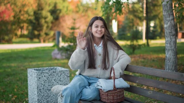 Close up of young woman sitting on wooden bench in outdoor park setting wearing casual clothing and interacting with items like woven basket. scene features natural daylight lighting green