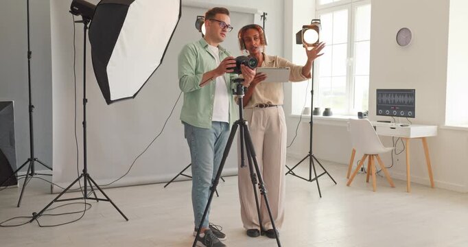 Photographers teamwork in studio review camera tablet. Wide studio shows colleagues planning a shoot and reviewing frames on tablet by camera and tripod. Clear teamwork and studio workflow.