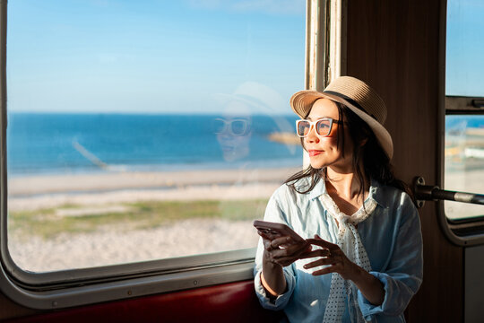 Young female traveler holding a mobile phone and enjoying the sea view while traveling by train during her summer vacation