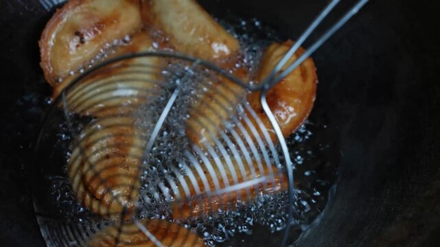 Slices of food frying in hot oil with a metal skimmer in a dark pan, showing bubbling oil and cooking process in close up
