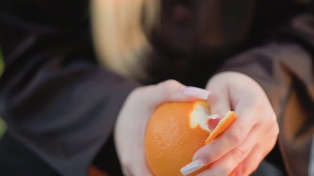 Close up of woman with long blonde hair carefully peeling orange revealing its texture and segments. Her hands display well manicured nails as woman interacts with fruit in soft lighting