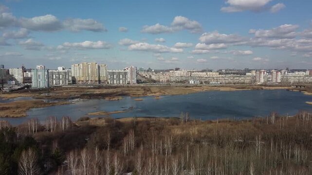 Blue sky with fluffy clouds above a quiet lake. Tall buildings stretch behind the water's edge. Dry grass and trees frame the natural landscape. Urban and wild coexist in serene balance